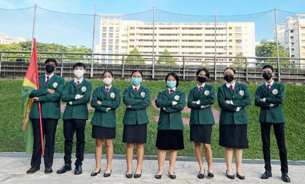 Seven students in green blazers and face masks stand with arms crossed outdoors near a flag and building.